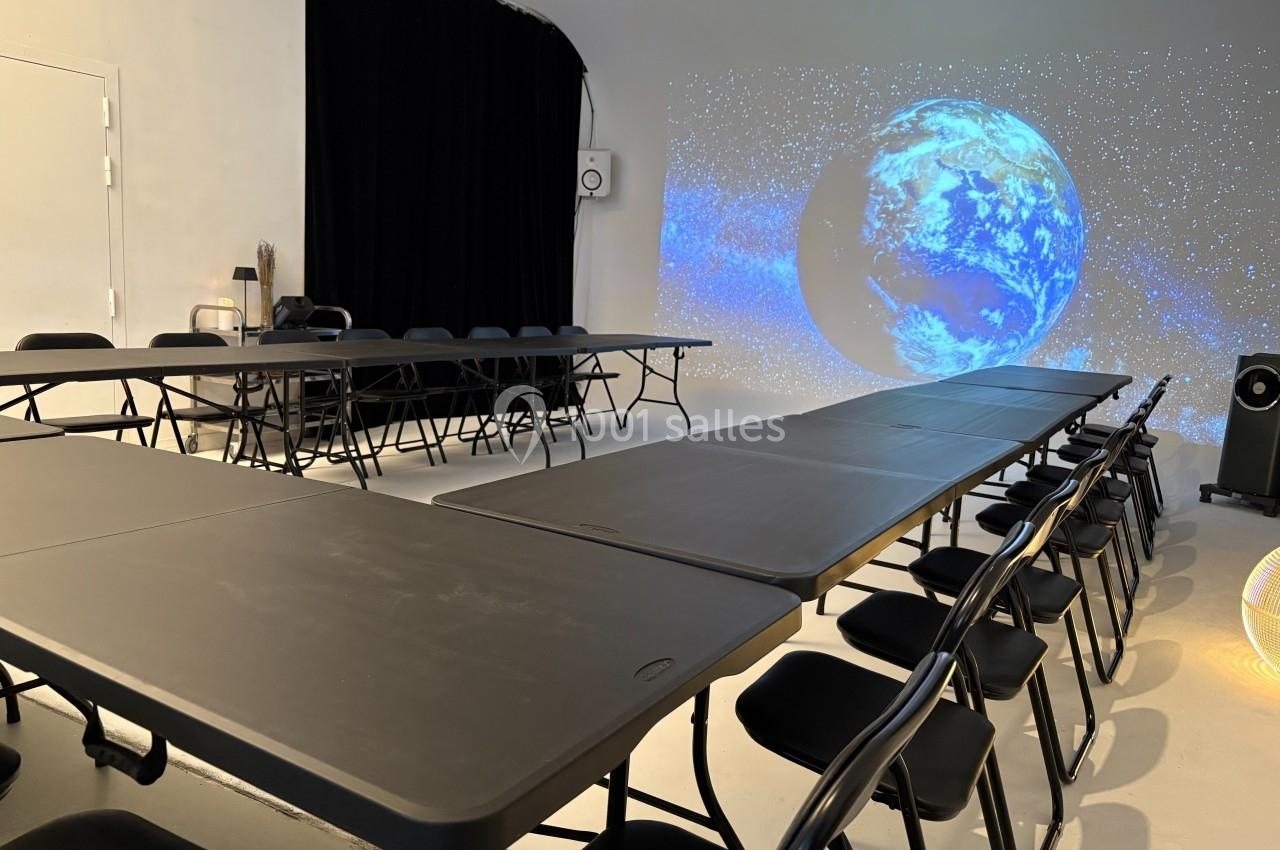 Salle avec des tables et chaises noires disposées en U, écran projetant une image de la Terre sur un fond étoilé.