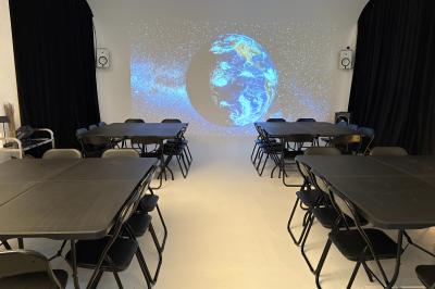 Salle avec des tables et chaises noires disposées en U, projection d’un paon coloré sur un mur blanc, rideaux noirs à droite.