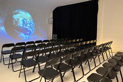 Salle avec des tables et chaises noires disposées en U, projection d’un paon coloré sur un mur blanc, rideaux noirs à droite.