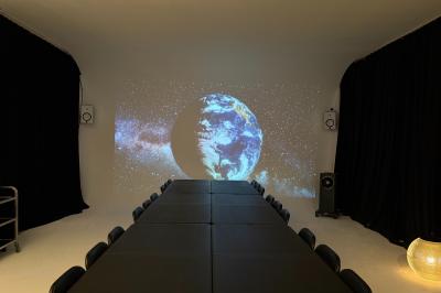 Salle avec des tables et chaises noires disposées en U, projection d’un paon coloré sur un mur blanc, rideaux noirs à droite.