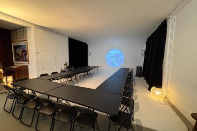 Salle avec des tables et chaises noires disposées en U, projection d’un paon coloré sur un mur blanc, rideaux noirs à droite.