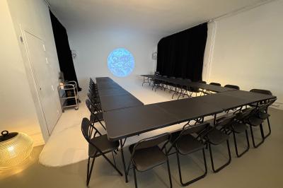 Salle avec des tables et chaises noires disposées en U, projection d’un paon coloré sur un mur blanc, rideaux noirs à droite.