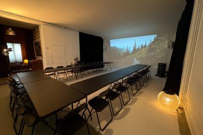 Salle avec des tables et chaises noires disposées en U, projection d’un paon coloré sur un mur blanc, rideaux noirs à droite.
