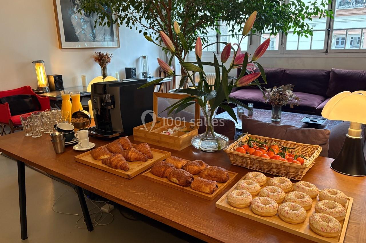 Table avec viennoiseries, fruits, boissons et machine à café dans un espace lumineux avec canapé et plantes.