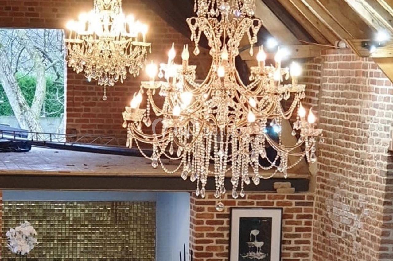 Deux lustres en cristal suspendus dans une pièce aux murs en briques, avec une grande fenêtre et un plafond en bois.