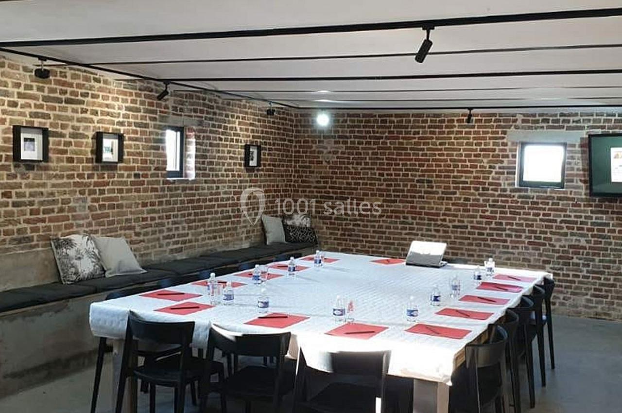 Salle de réunion avec murs en briques, table rectangulaire dressée avec documents rouges, chaises noires et lumière…