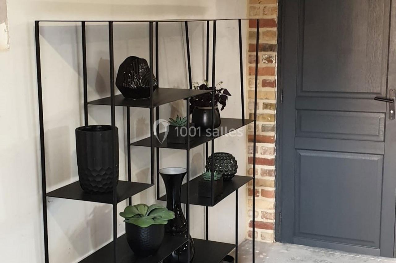 Étagère noire moderne avec des objets décoratifs et des plantes, placée près d'un mur en brique et d'une porte grise.