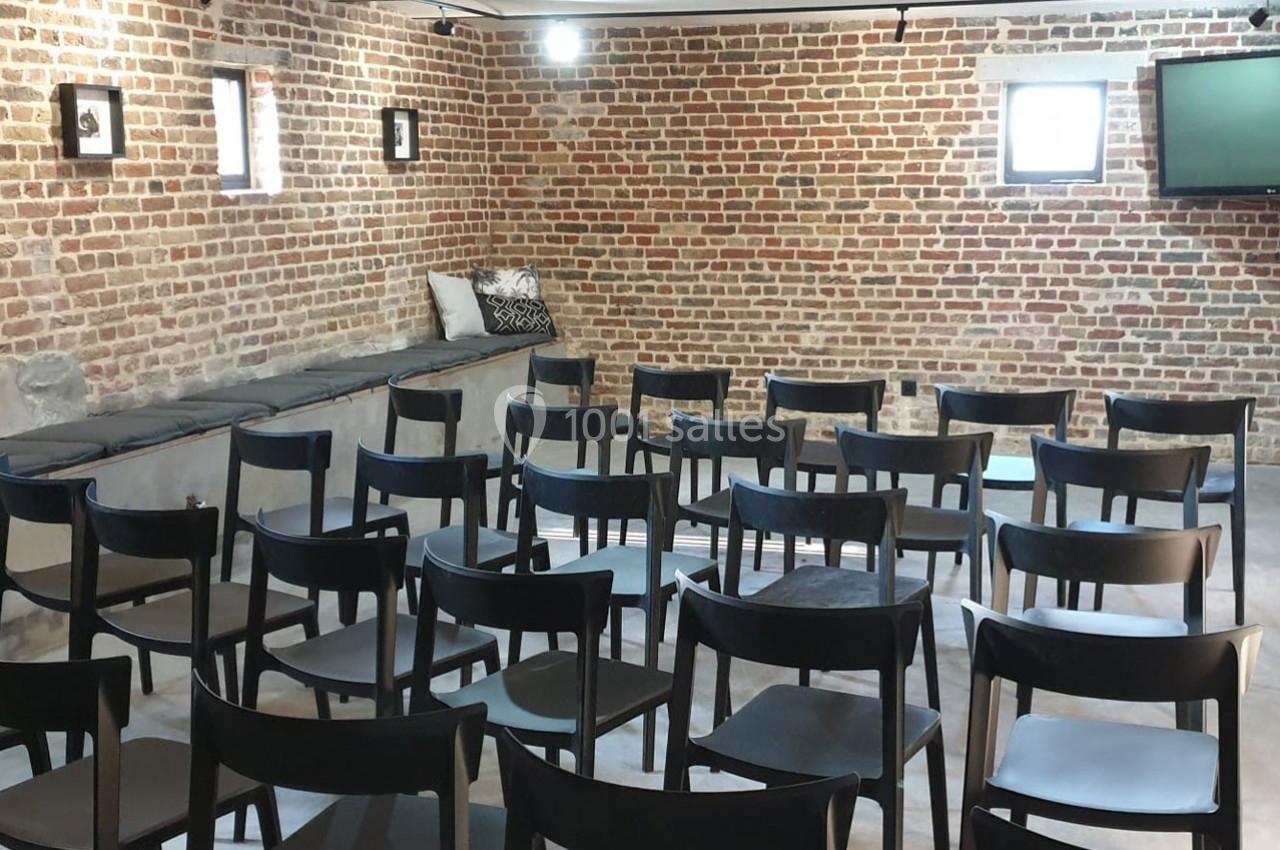 Salle avec des murs en briques, rangées de chaises noires alignées et un écran fixé au mur.