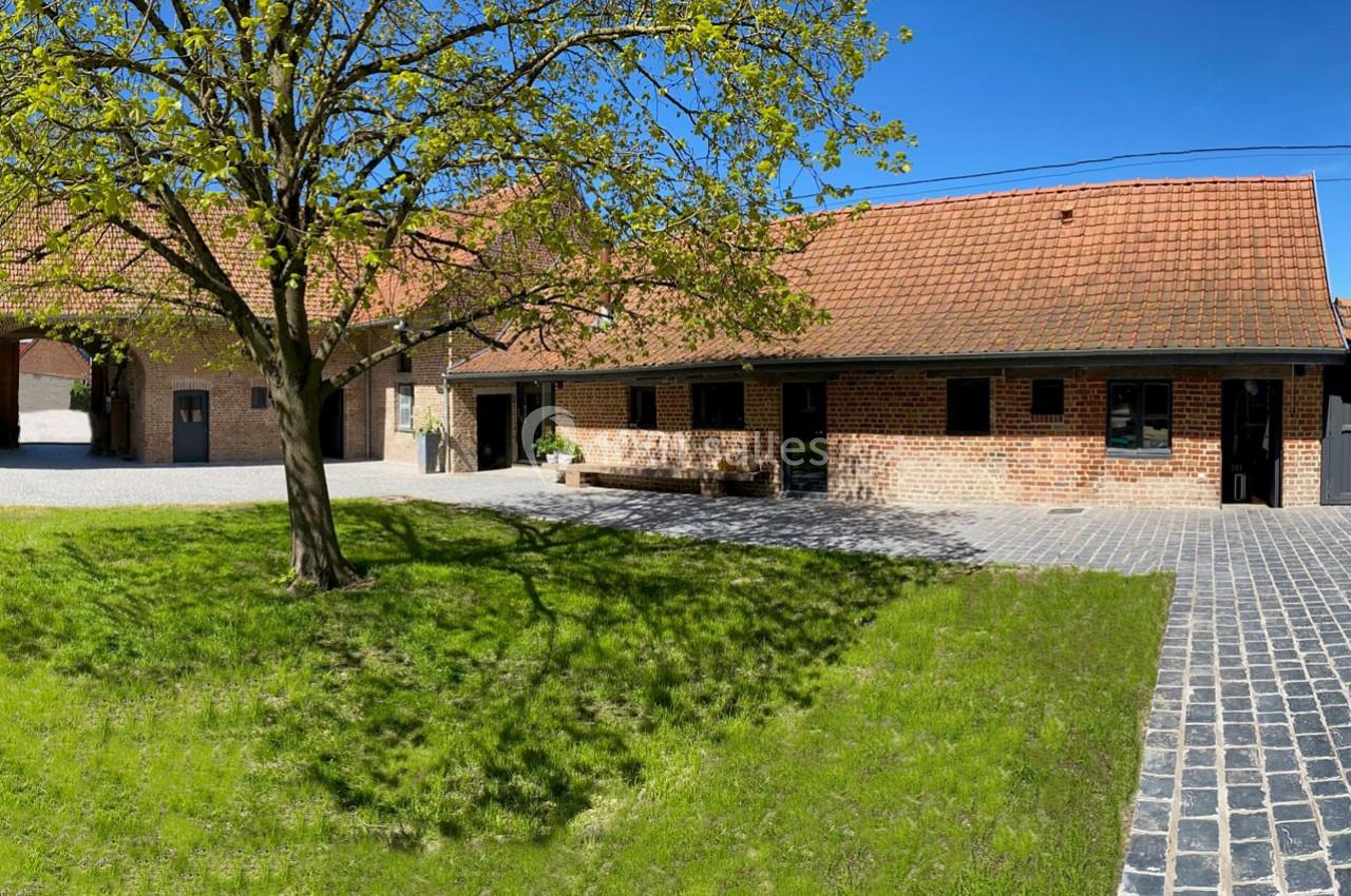 Bâtiment en briques rouges avec toit en tuiles, cour pavée, pelouse et arbre sous un ciel bleu.