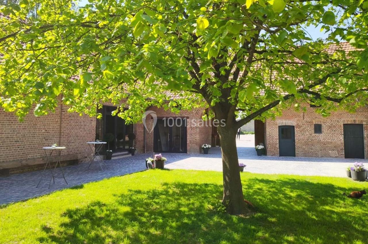 Cour pavée avec un arbre au centre, entourée de bâtiments en briques et de pots de fleurs sous un ciel ensoleillé.