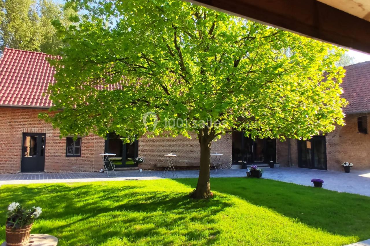 Cour ensoleillée avec un arbre central, entourée de bâtiments en briques et de mobilier de jardin.