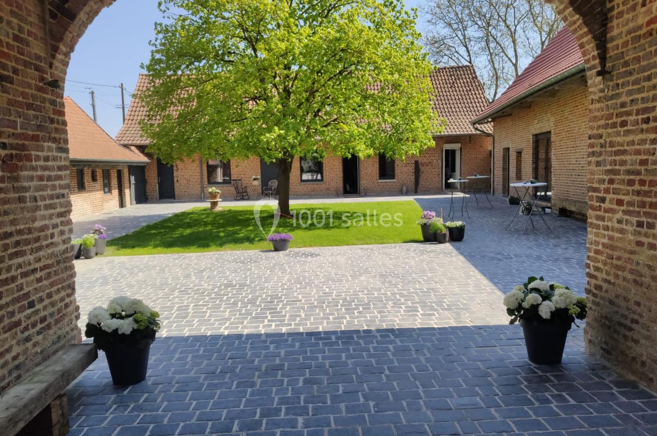 Cour pavée avec un arbre central, entourée de bâtiments en briques, pots de fleurs et mobilier de jardin.