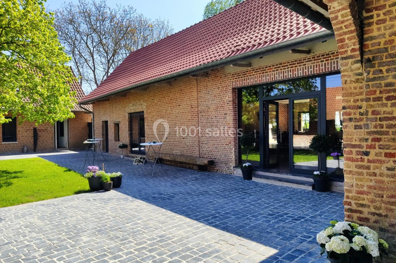 Cour pavée avec bâtiment en briques rouges, grandes fenêtres et plantes en pots, entourée de verdure.