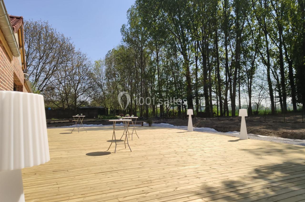 Terrasse en bois avec quelques tables et lampes, bordée d'arbres sous un ciel dégagé.