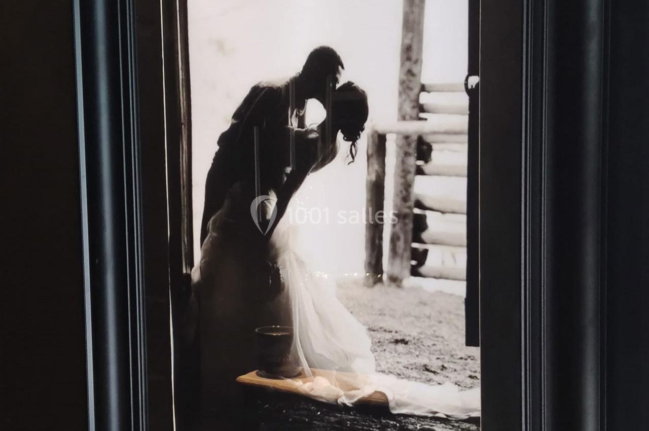 Photographie en noir et blanc d'un couple s'embrassant, la femme en robe de mariée, devant une structure en bois.