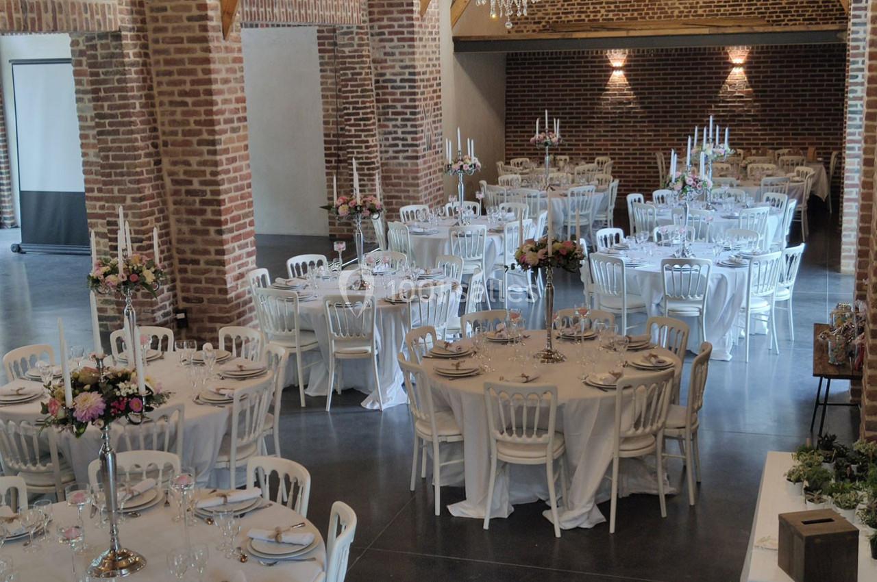 Salle de réception avec tables rondes dressées, nappes blanches, chandeliers et décorations florales.