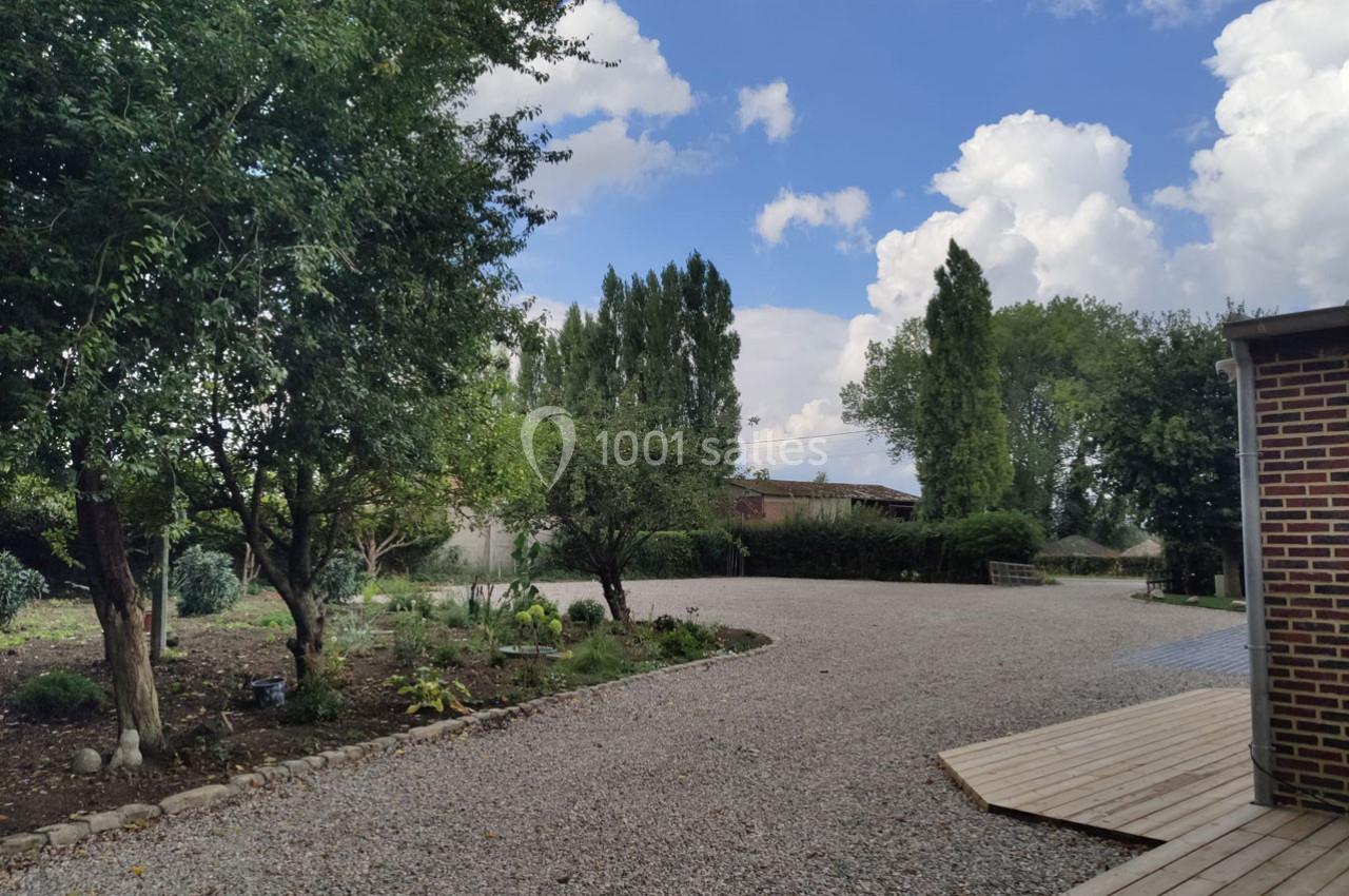 Cour extérieure avec allée en gravier, bordée d'arbres et d'arbustes, et terrasse en bois à droite sous un ciel…