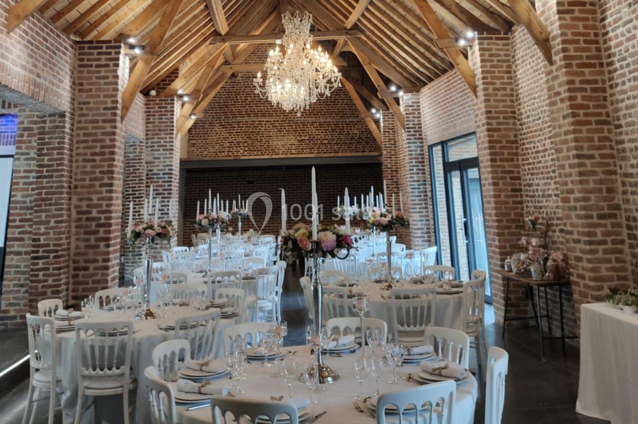 Salle de réception avec murs en briques, poutres apparentes, tables rondes dressées et lustre central.