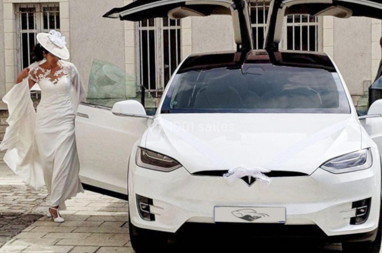 Photo David J. Location #2 Une mariée en robe blanche sort d'une voiture électrique blanche décorée pour un mariage, avec des portes papillon ouvertes.