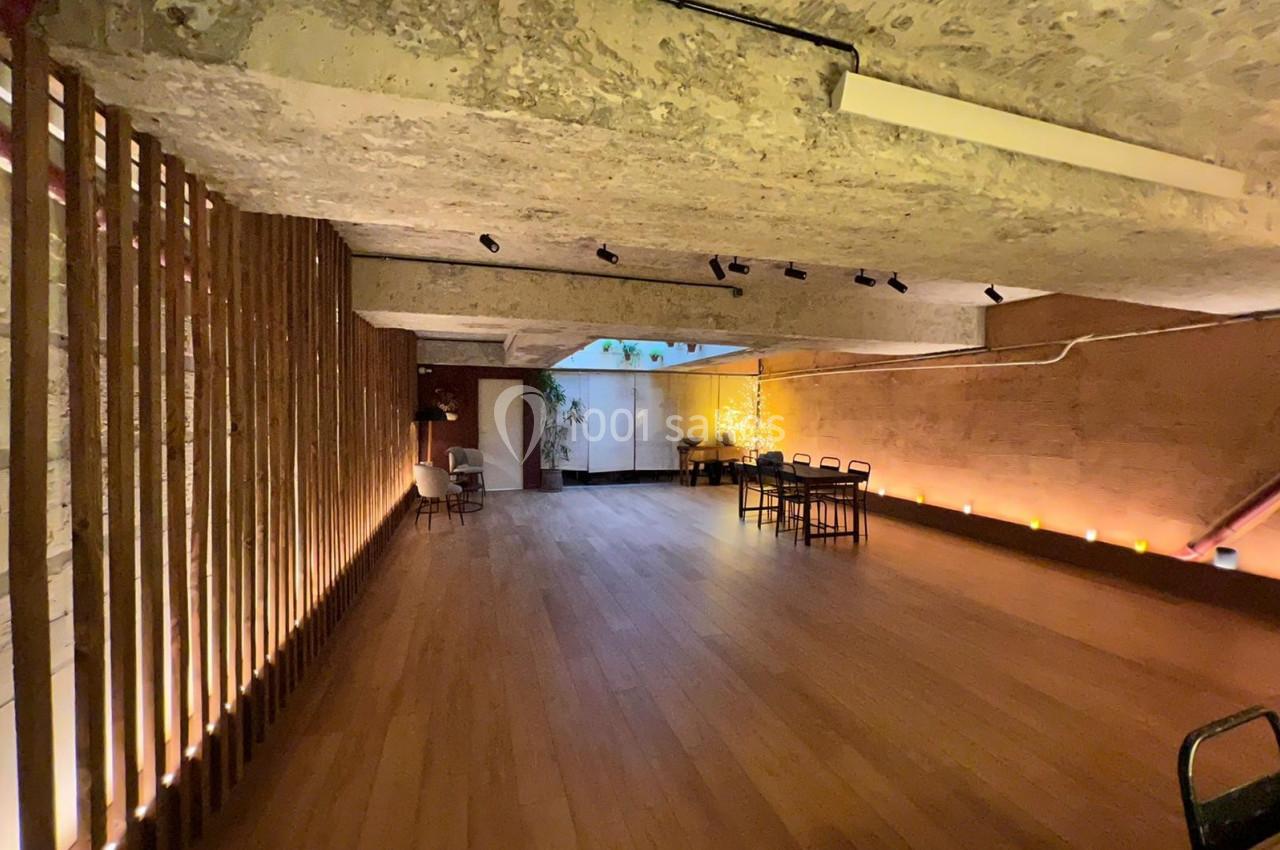 Salle avec parquet en bois, murs en pierre et éclairage tamisé, aménagée avec quelques tables et chaises.