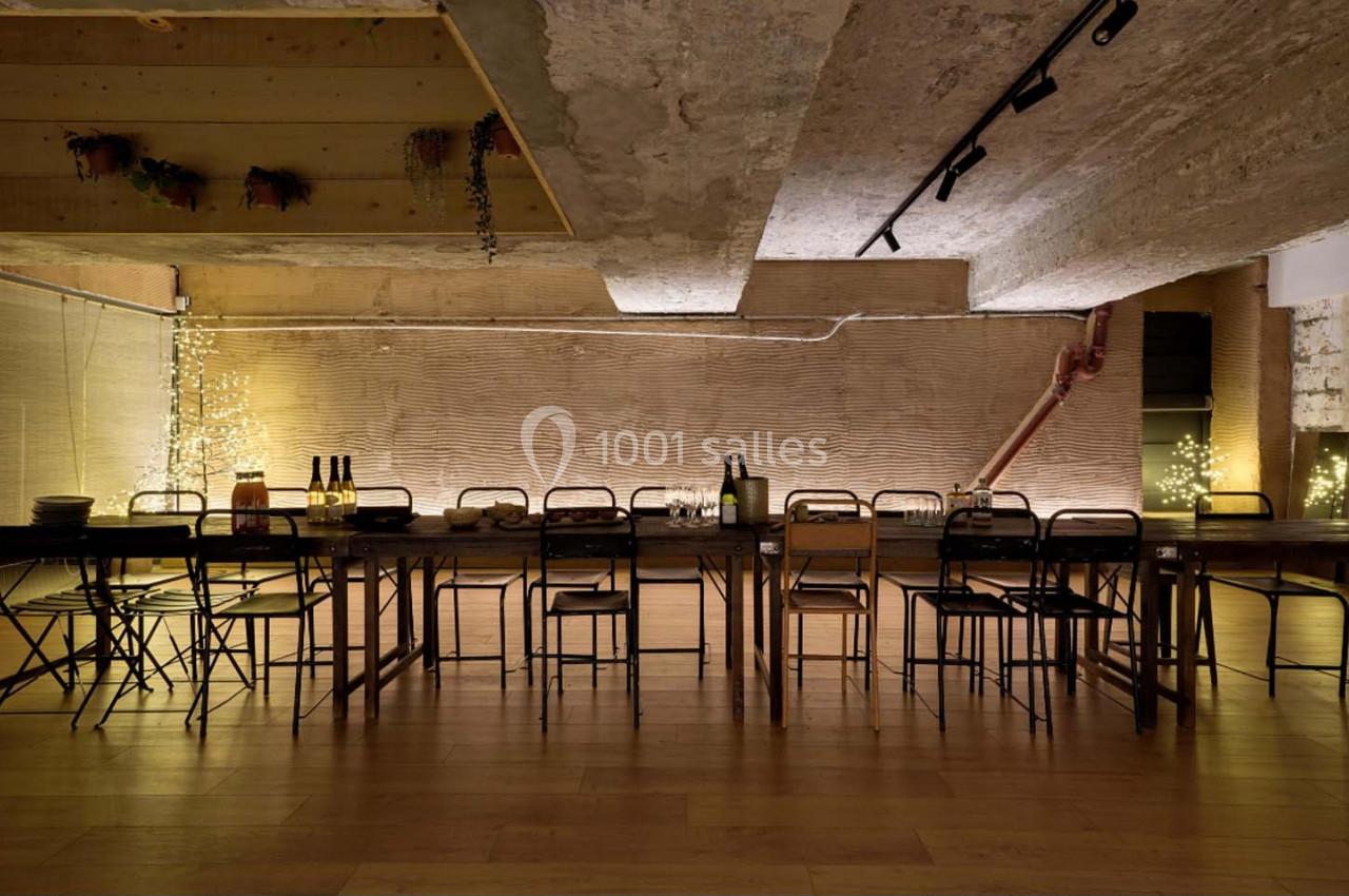 Grande table en bois entourée de chaises dans une pièce au style industriel avec éclairage tamisé et murs texturés.