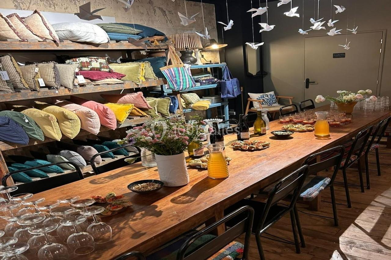 Table en bois dressée avec boissons, fleurs et collations, entourée d'étagères garnies de coussins colorés et décorations.