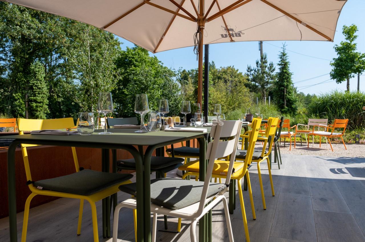 Terrasse ensoleillée avec tables et chaises colorées, entourée de verdure et protégée par un grand parasol beige.