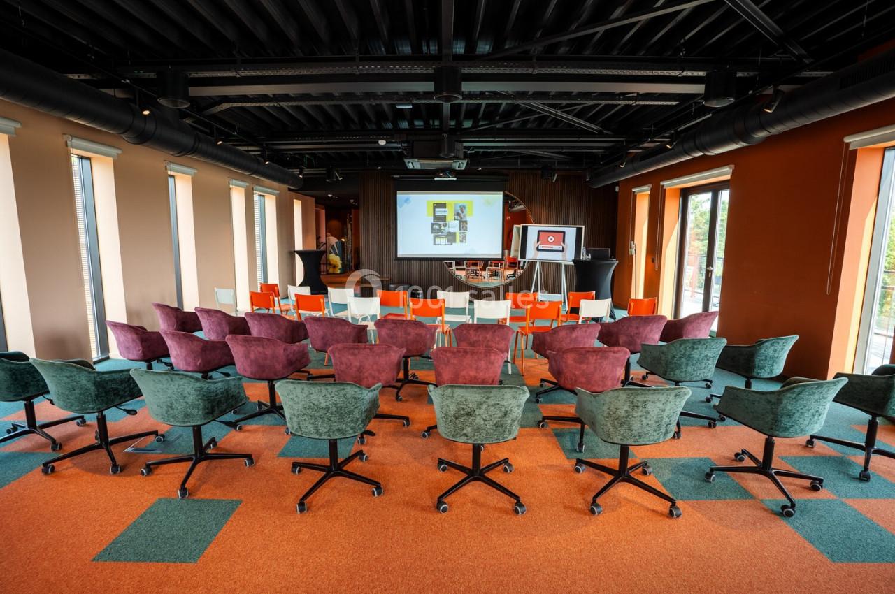 Salle de conférence vide avec des rangées de chaises colorées face à un écran de présentation.
