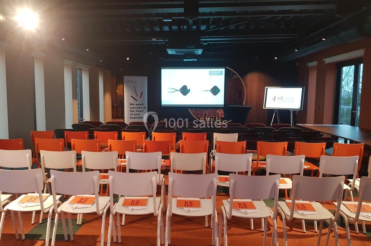Salle de conférence vide avec des rangées de chaises blanches et orange, écran de présentation et supports sur les sièges.