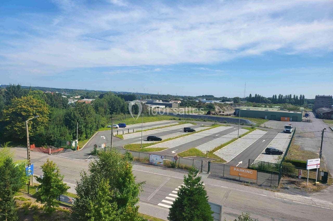 Vue aérienne d'un parking vide entouré de végétation, avec quelques voitures stationnées et un ciel dégagé.