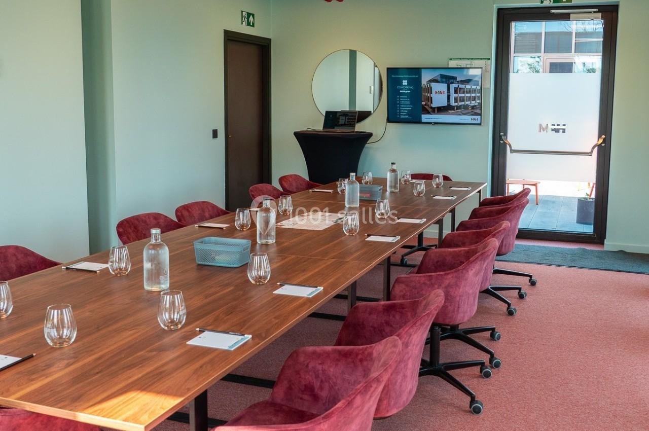 Salle de réunion lumineuse avec une grande table en bois, chaises rouges, écran et porte vitrée donnant sur l'extérieur.