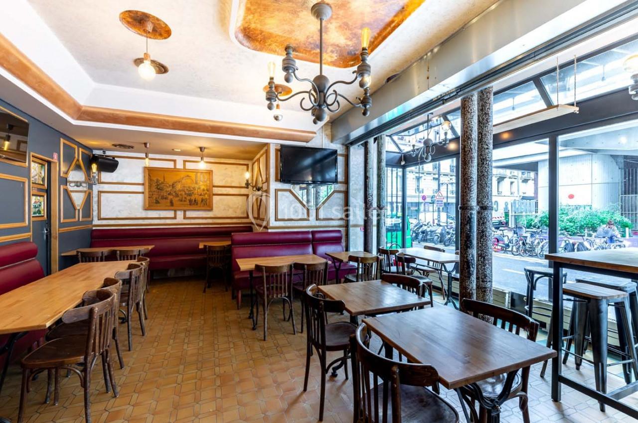 Salle de restaurant lumineuse avec tables en bois, banquettes rouges, lustres et grande baie vitrée donnant sur la rue.