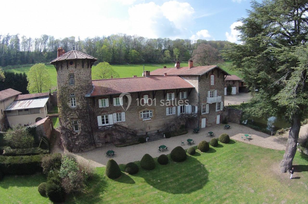 Manoir en pierre entouré de jardins, avec une tour, des tables extérieures et une forêt en arrière-plan.
