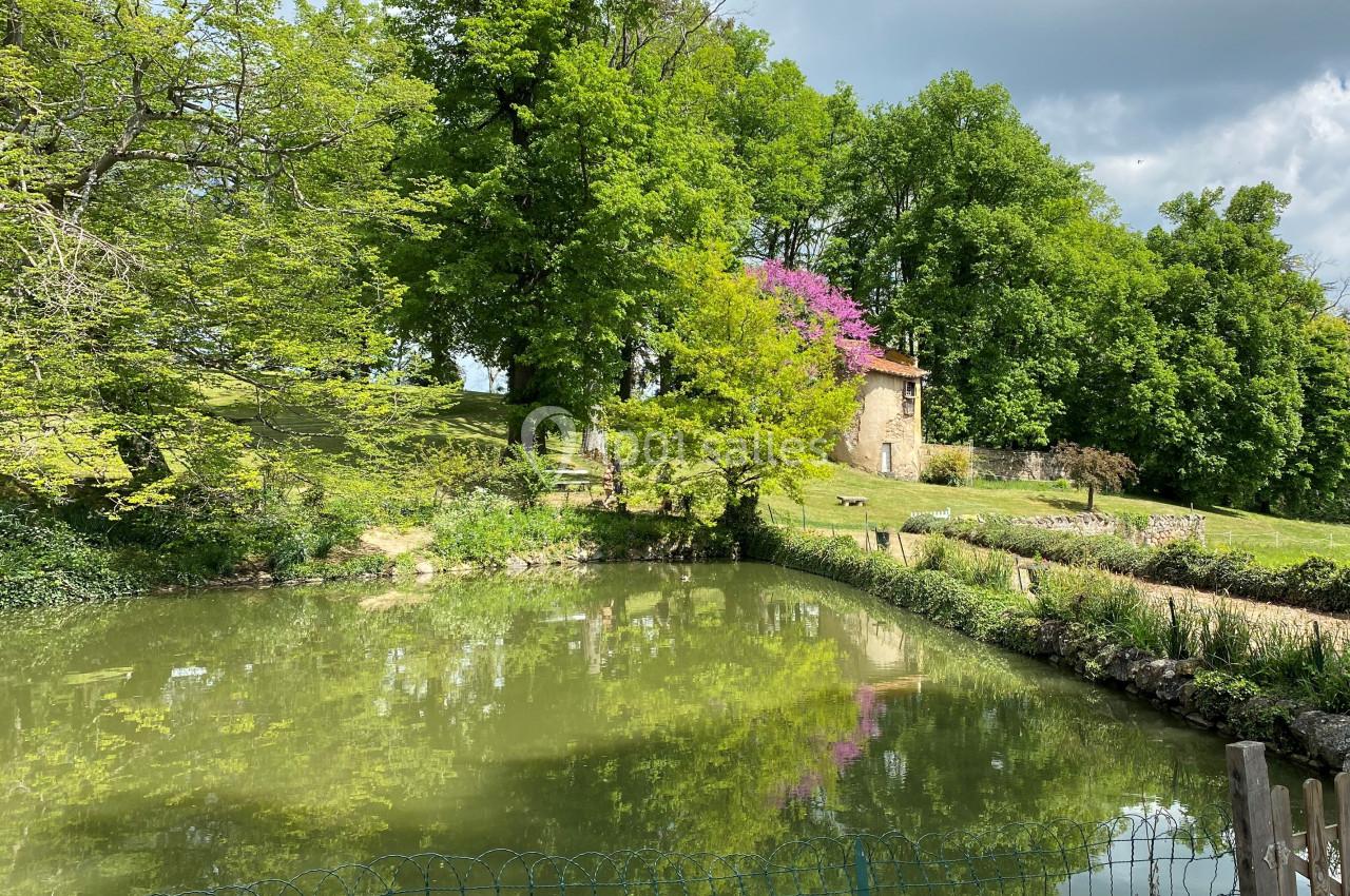 Petit étang entouré de verdure avec un bâtiment en pierre et des arbres fleuris en arrière-plan.