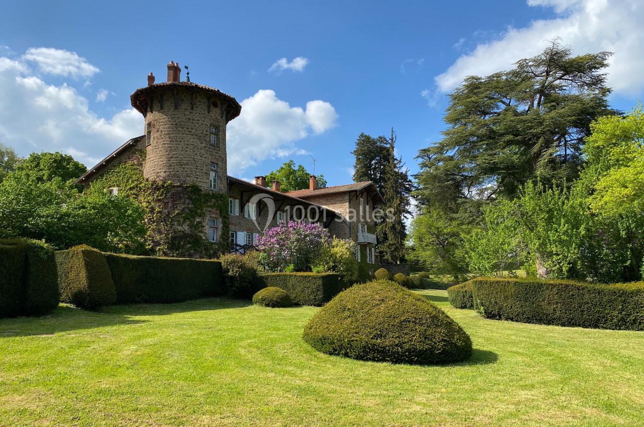 Grande maison en pierre avec une tour ronde, entourée de haies taillées, d'arbres et d'une pelouse sous un ciel bleu.