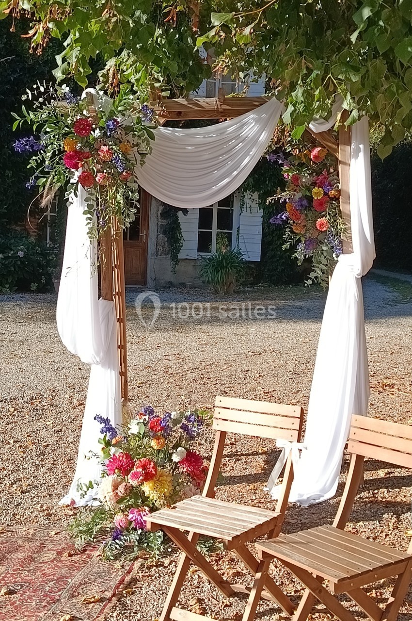 Arche de mariage en bois décorée de fleurs colorées et de voilages blancs, avec deux chaises en bois devant.