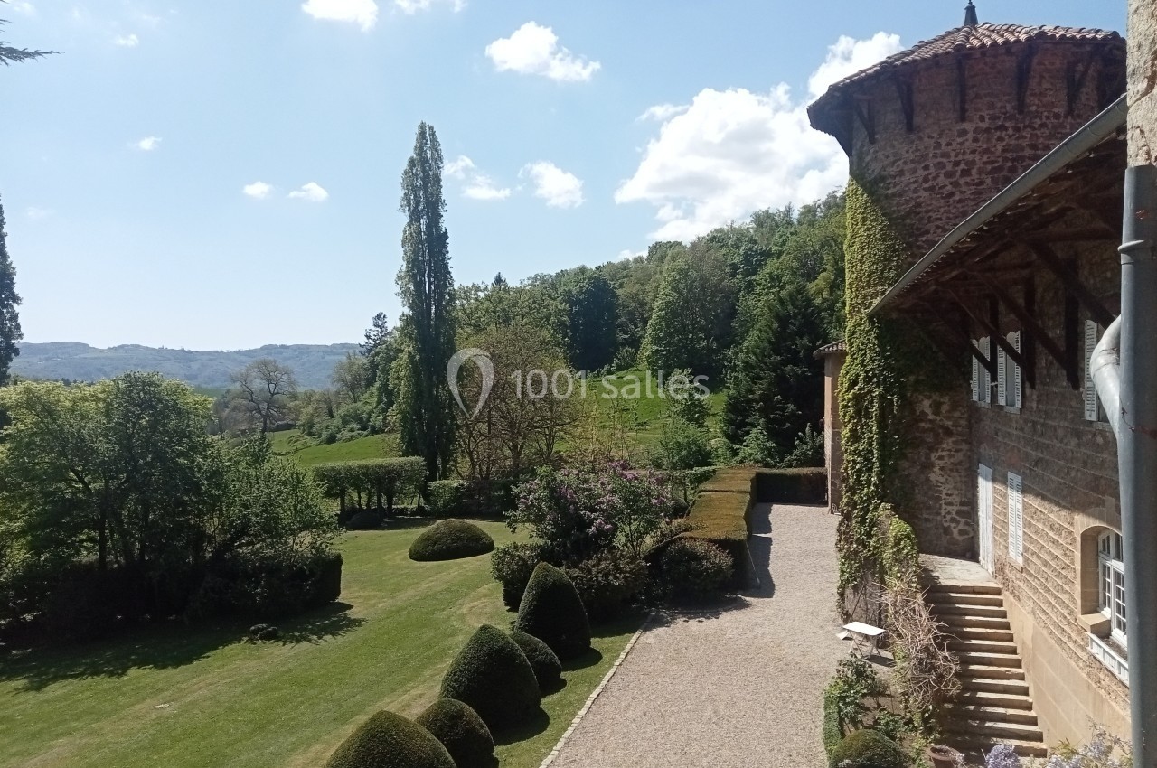 Vue d'un jardin paysager avec pelouse, arbustes taillés, arbres et un bâtiment en pierre sous un ciel ensoleillé.
