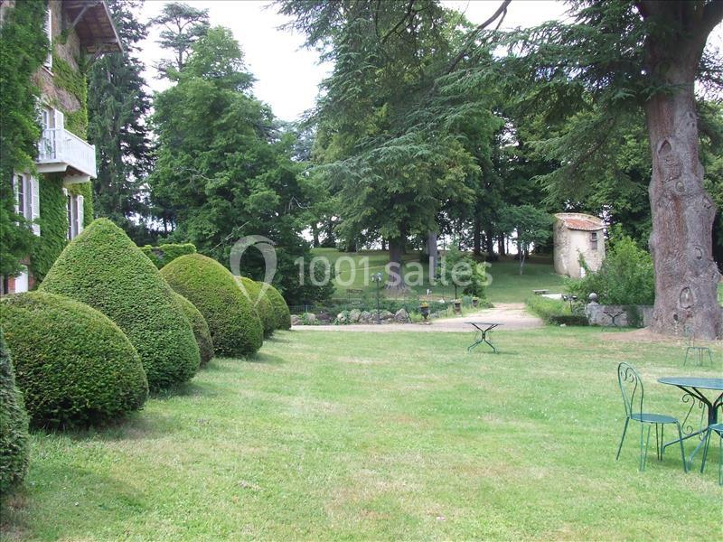 Jardin verdoyant avec pelouse, arbustes taillés, grands arbres, petite bâtisse en pierre et mobilier de jardin en métal.