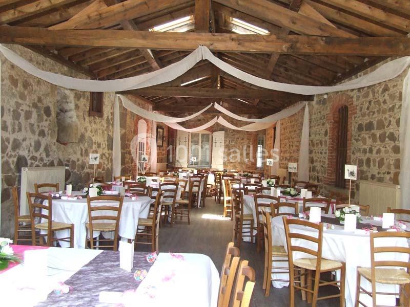 Salle en pierre avec charpente apparente, tables rondes dressées pour un événement, décorations sobres et lumineuses.