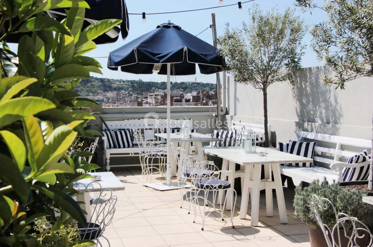 Terrasse ensoleillée avec tables blanches, chaises en métal, parasols bleus et vue sur une ville en arrière-plan.