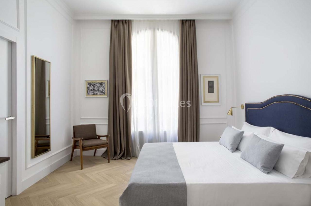 Chambre lumineuse avec lit double, tête de lit bleue, rideaux beige, chaise en bois et parquet clair.