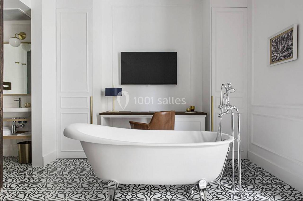 Salle de bain lumineuse avec baignoire îlot, sol à motifs, bureau en bois, fauteuil en cuir et télévision murale.