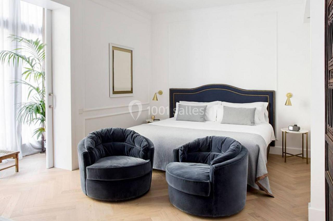 Chambre lumineuse avec lit double, fauteuils bleus, parquet clair, grande fenêtre et décoration minimaliste.