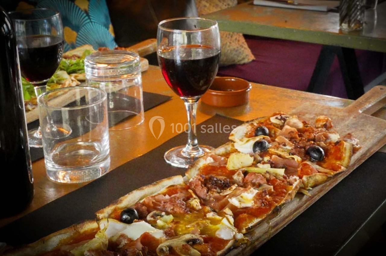 Planche de pizza garnie de charcuterie, olives et fromage, servie avec un verre de vin rouge sur une table en bois.