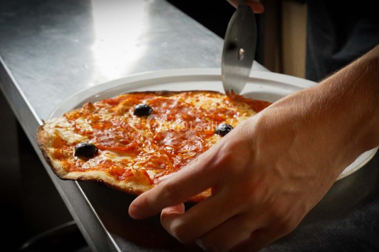 Une main découpe une pizza fine garnie de fromage, sauce tomate et olives noires à l'aide d'une roulette à pizza.