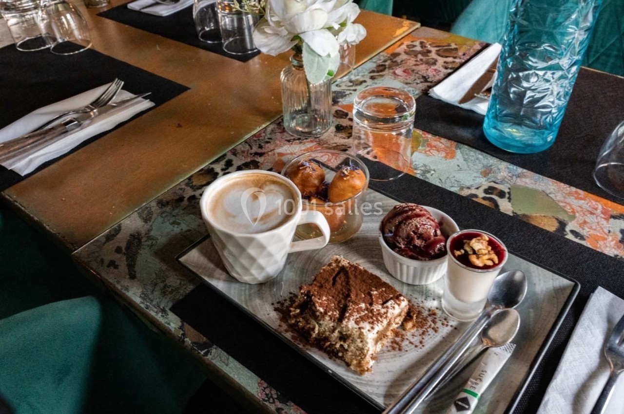 Assiette avec tiramisu, glace, beignets et cappuccino sur une table décorée, avec couverts et fleurs en arrière-plan.
