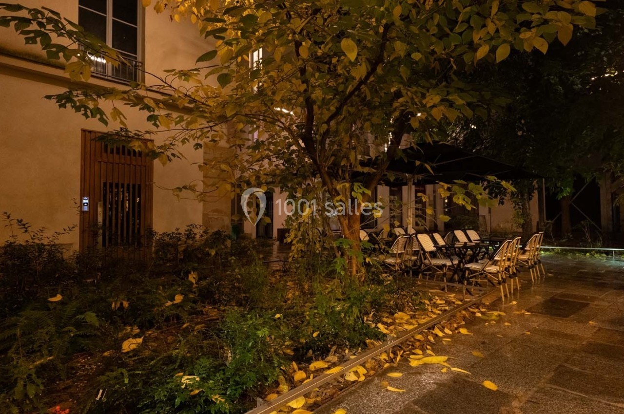 Cour intérieure éclairée de nuit, avec des chaises et tables sous un arbre aux feuilles jaunies en automne.
