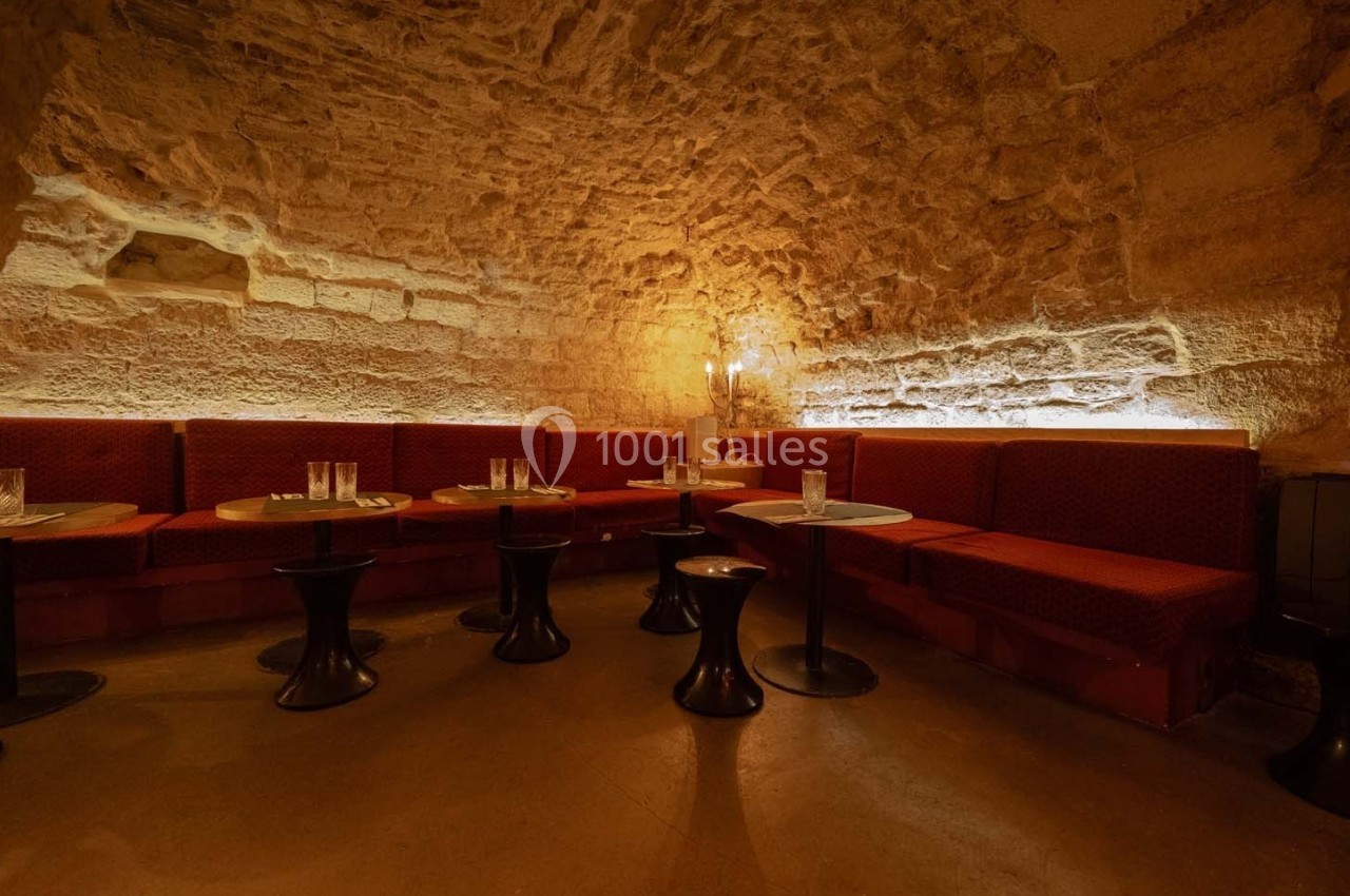 Salle voûtée en pierre avec banquettes rouges, petites tables rondes et éclairage tamisé.