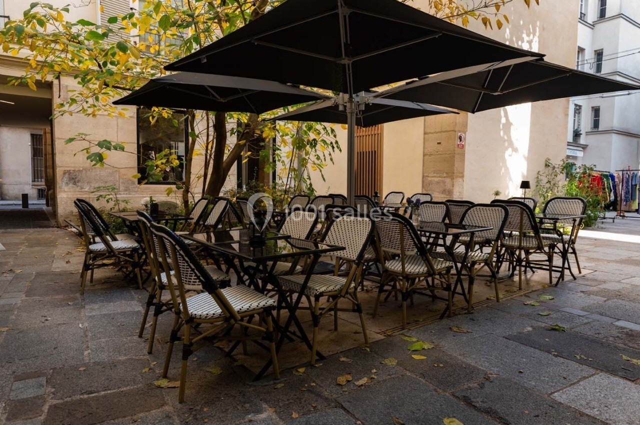 Terrasse extérieure avec tables et chaises en osier sous des parasols noirs, entourée d'arbres et de bâtiments.