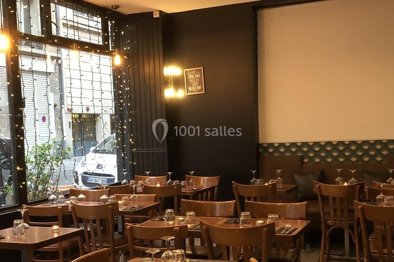 Salle de restaurant avec tables en bois, chaises assorties, grandes fenêtres et guirlandes lumineuses.